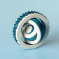 May include: An abstract sculpture with a white circular frame and a textured, teal edge. Inside, teal and white ribbons create a swirling pattern. The sculpture is set against a light blue background. The sculpture is approximately 10 cm in diameter.