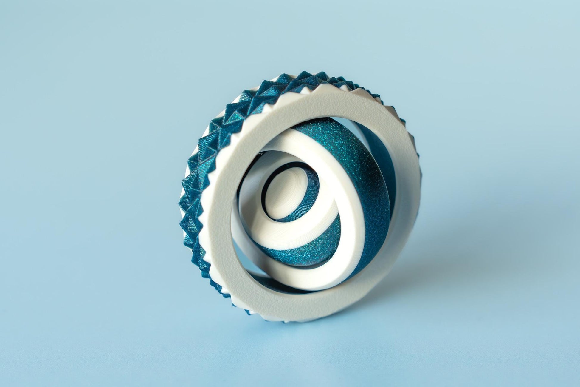May include: An abstract sculpture with a white circular frame and a textured, teal edge. Inside, teal and white ribbons create a swirling pattern. The sculpture is set against a light blue background. The sculpture is approximately 10 cm in diameter.