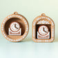 May include: Two decorative clocks in a light brown colour, one round and one arched, each housing a small clock with a white face and brown hands. The clock faces are surrounded by a brown frame with white floral accents. The clocks are on a light green background.