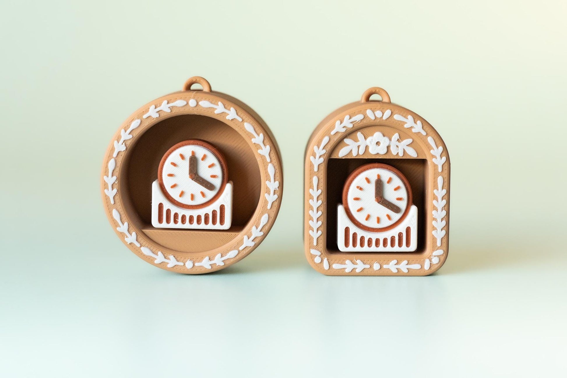 May include: Two decorative clocks in a light brown colour, one round and one arched, each housing a small clock with a white face and brown hands. The clock faces are surrounded by a brown frame with white floral accents. The clocks are on a light green background.